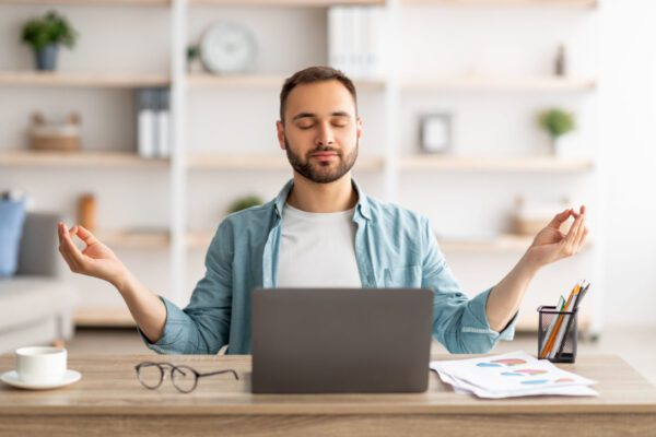 Workplace stress management. Calm Caucasian man meditating with closed eyes in front of laptop pc at home office. Young male freelancer feeling peaceful and balanced, doing yoga at his desk