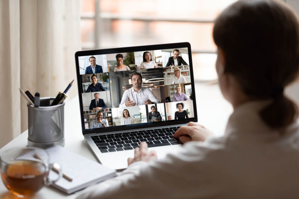 View over businesslady shoulder seated at workplace desk look at computer screen where collage of many diverse people involved at video conference